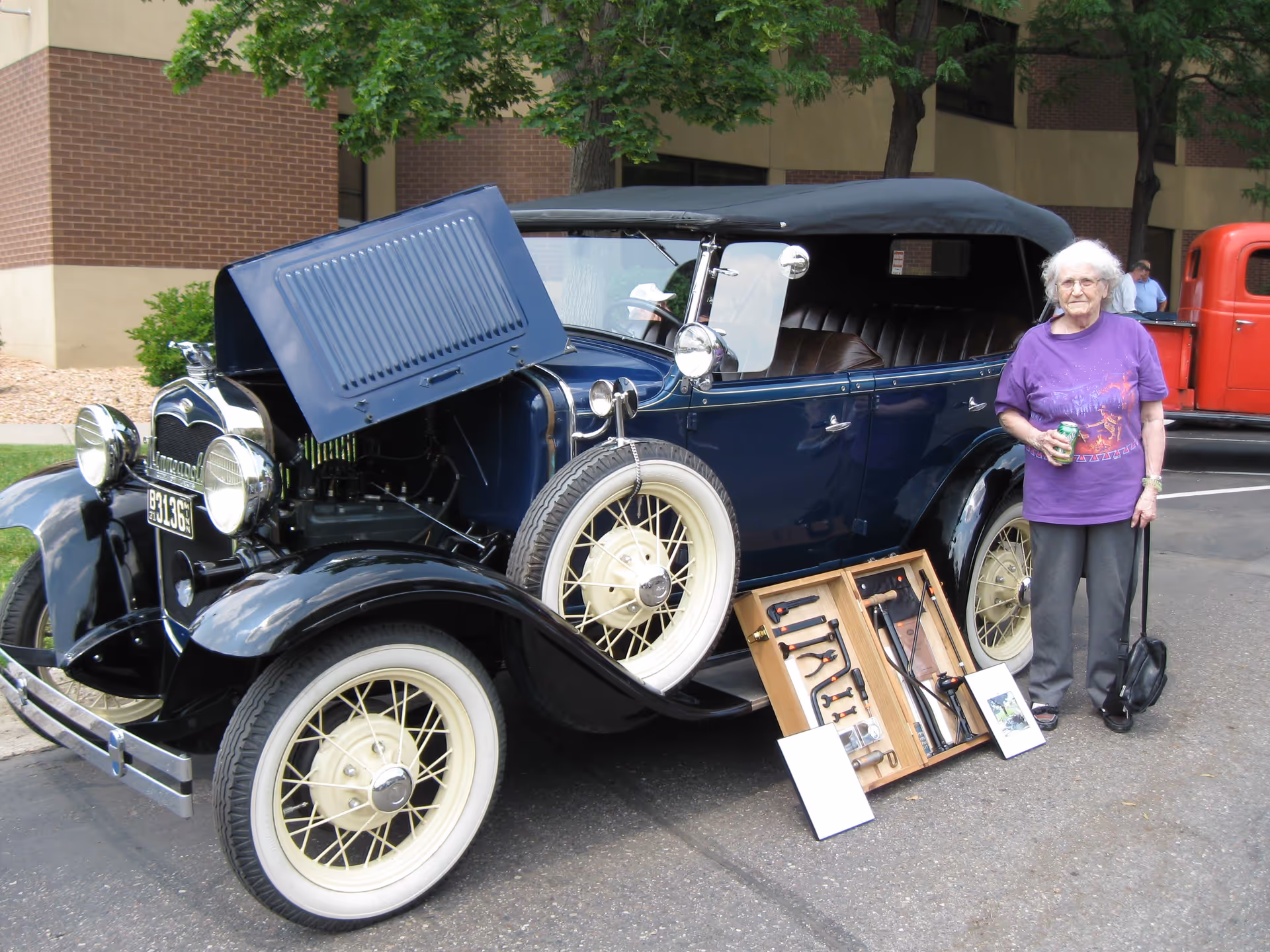 An elderly woman wearing a purple shirt and gray pants stands next to a vintage blue car with its hood open. A display board with various tools is propped against the car's front tire. The scene is outdoors in a parking lot with a brick building and trees in the background.