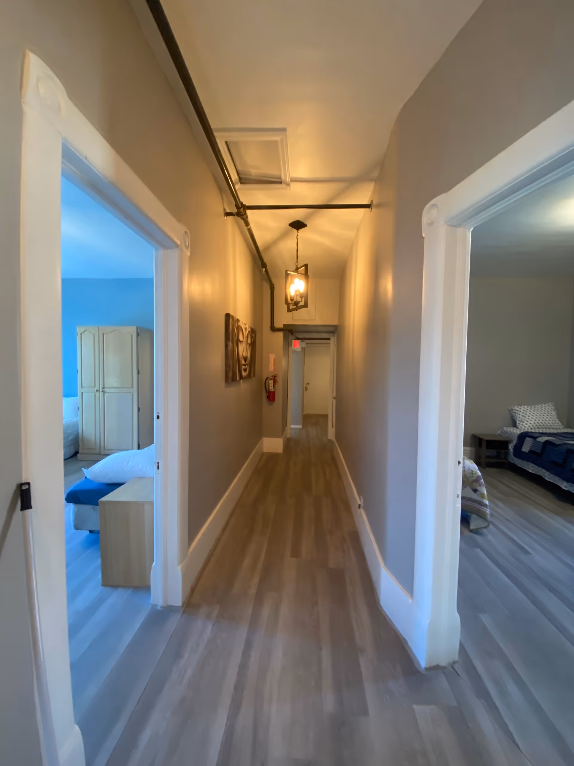 Long narrow hallway in a residential facility with bedrooms visible on both sides, wood-look floors, wall art, and a hanging light fixture.