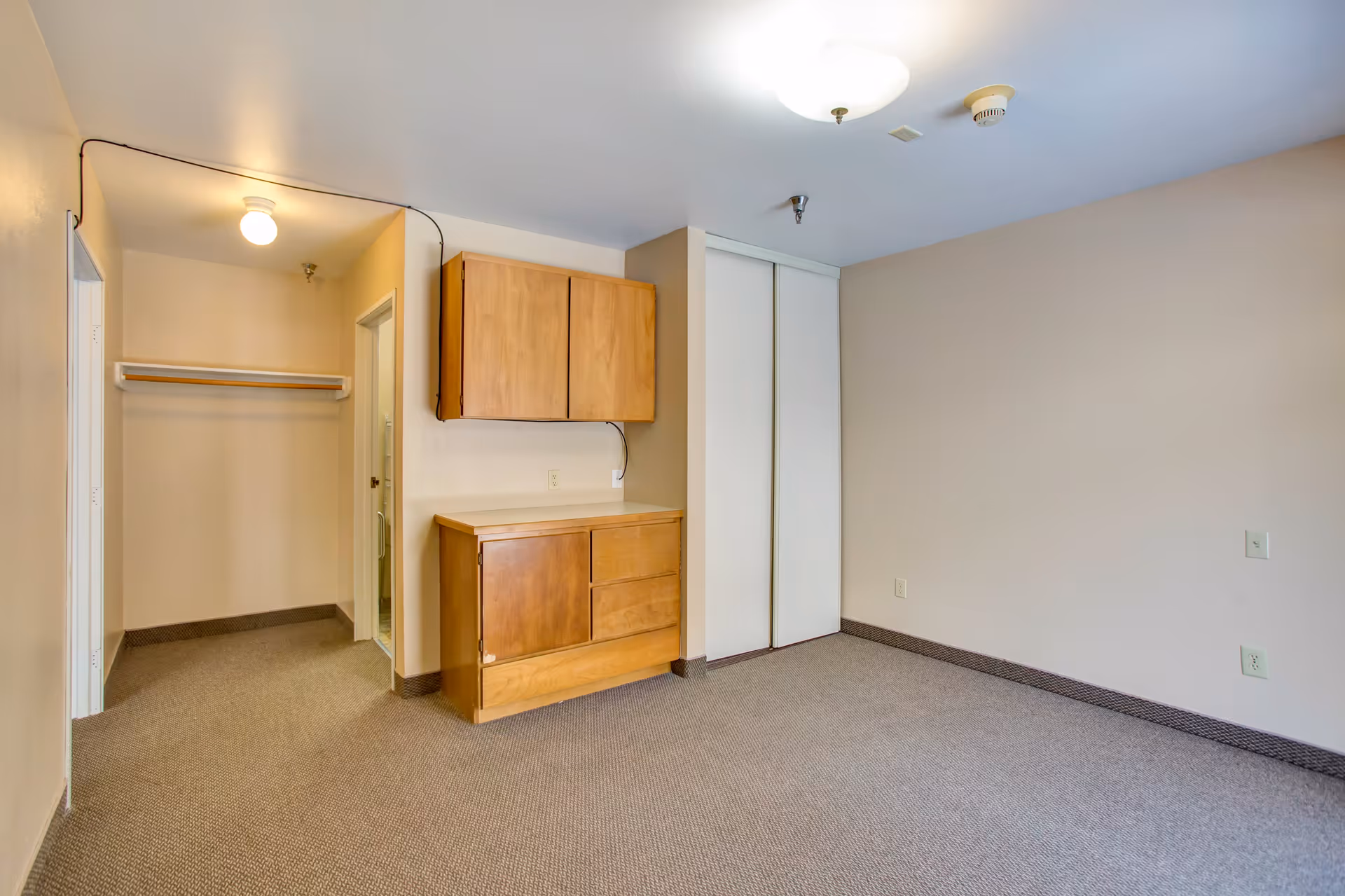 Empty room with beige walls and carpeted floor, featuring wooden cabinets mounted on the wall and below, a closet with sliding white doors, and an open doorway leading to a small walk-in closet with a hanging rod and a light fixture on the ceiling.