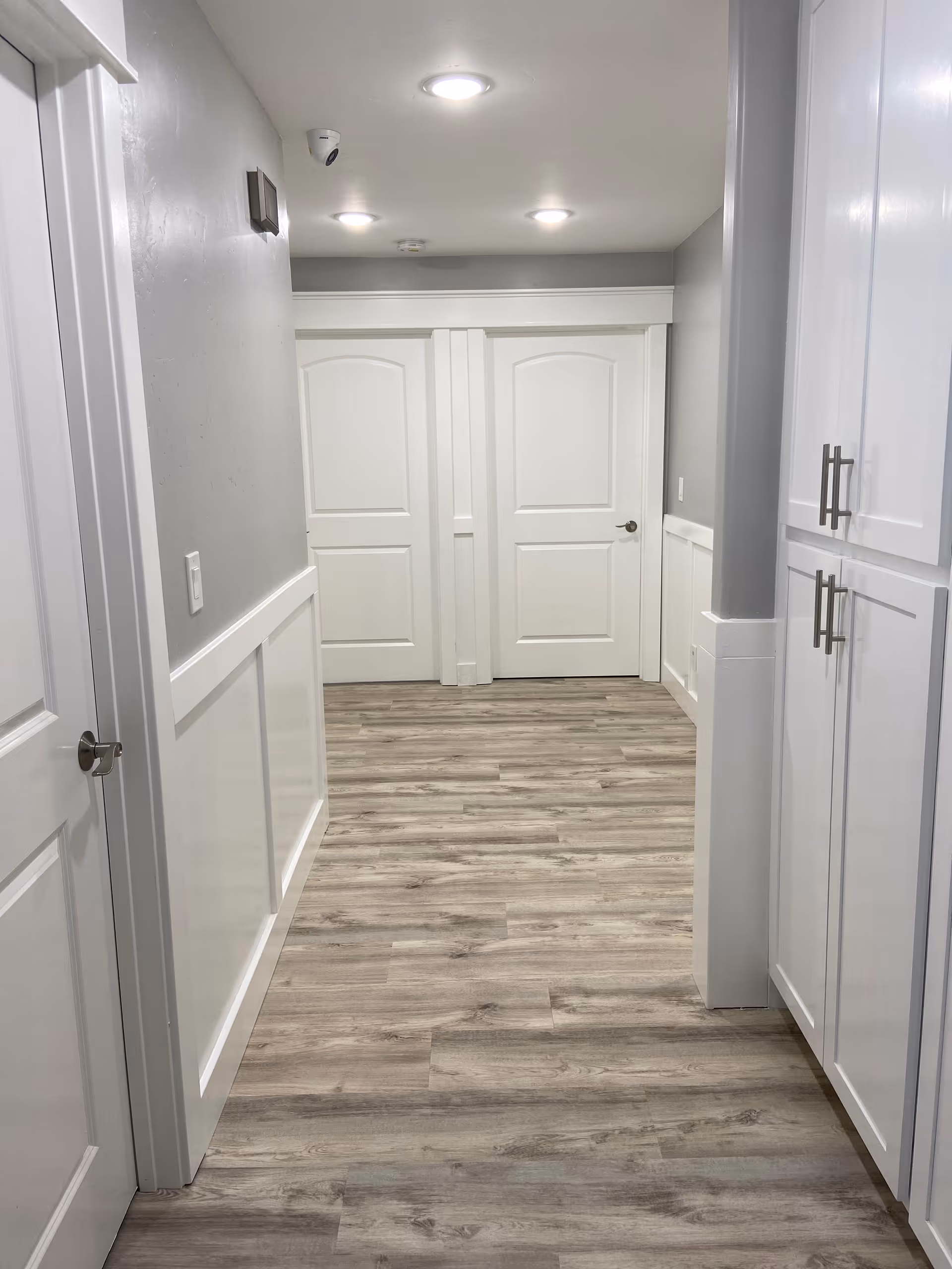 A clean, well-lit hallway with light gray walls and white trim. The floor is covered with wood-patterned vinyl or laminate flooring. There are three white doors visible, one on the left side and two at the end of the hallway. On the right side, there are white cabinets with silver handles. The ceiling has recessed lighting and a security camera is mounted on the left wall near the ceiling.