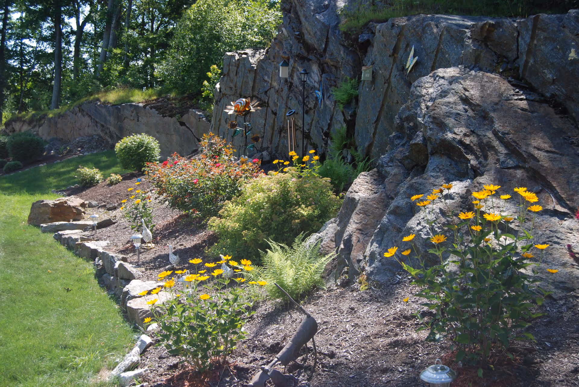 Sunlit landscaped rock garden with yellow flowers, shrubs, and birdhouses beside a grassy lawn.
