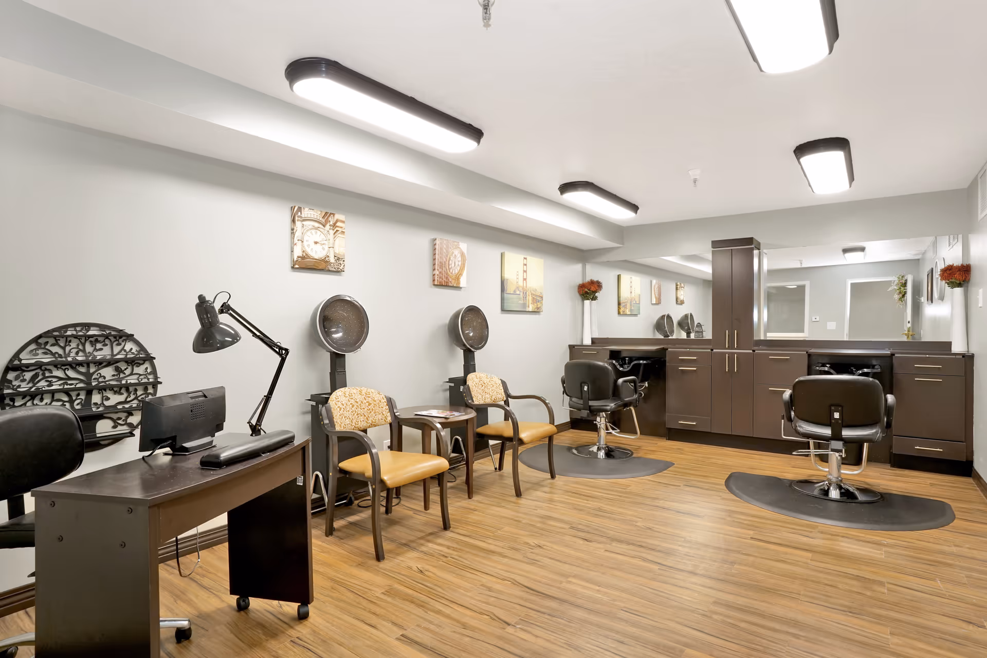 Interior view of a salon area with two hair drying chairs, two styling chairs in front of large mirrors, a desk with a lamp and computer, and two patterned chairs along the wall. The room has wood flooring, light gray walls, and ceiling lights. Decorative pictures and vases with flowers are on the walls and counters.