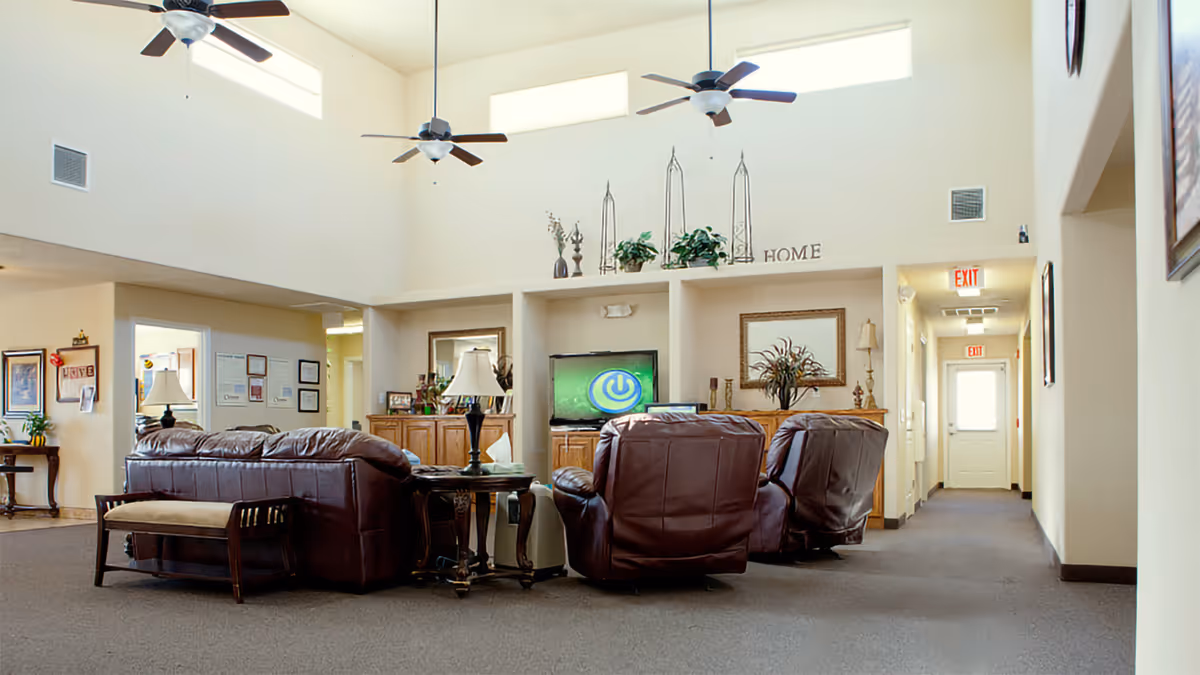A spacious living room area with high ceilings and ceiling fans. The room features brown leather sofas and armchairs arranged around a TV on a wooden cabinet. Decorative plants, lamps, and framed artwork adorn the room. There is a hallway with exit signs visible in the background.