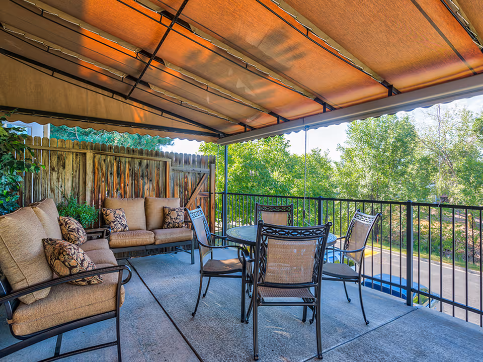 Covered outdoor patio area with cushioned seating including a sofa and chairs around a round table, surrounded by a black metal railing and overlooking a green landscape with trees.