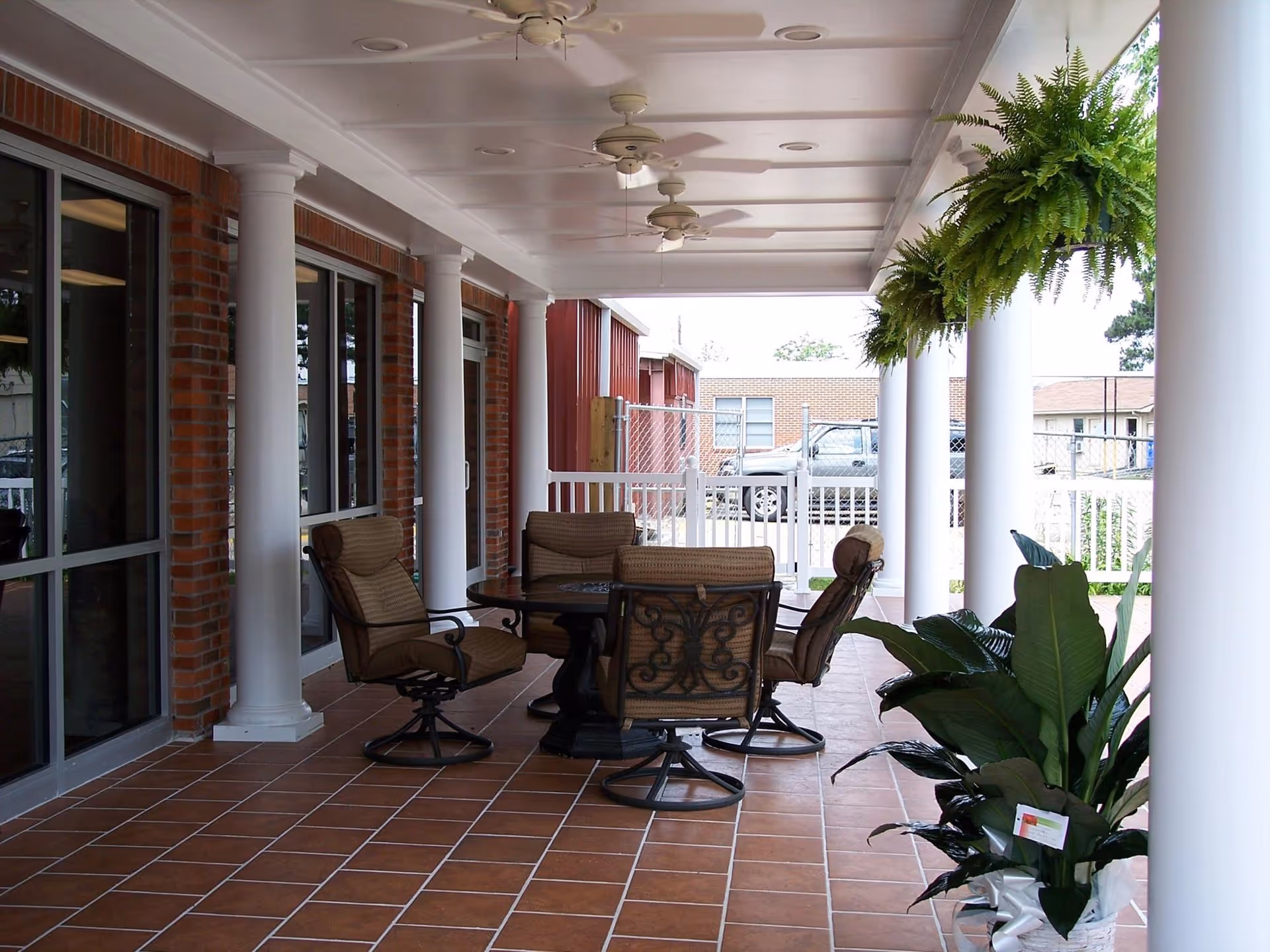 A covered outdoor patio with a round table and cushioned chairs, white columns, ceiling fans, hanging ferns, and potted plants.