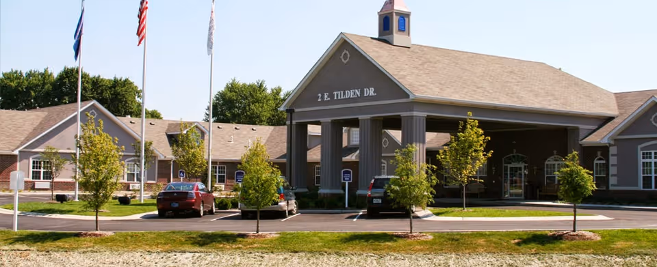 Exterior view of Brownsburg Meadows facility showing a large building with a covered entrance supported by columns. The building has a sign with the address '2 E. Tilden Dr.' There are three flagpoles with flags in front of the building, several small trees, and a parking area with cars.