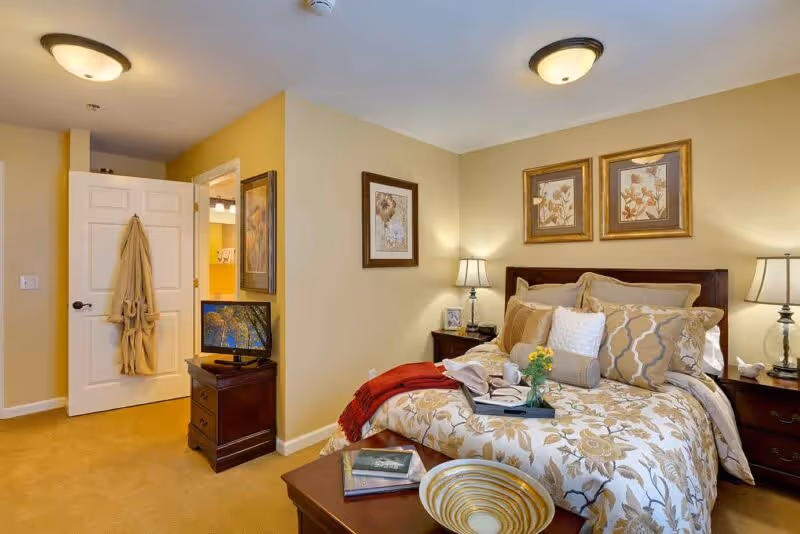 A cozy bedroom with a neatly made bed featuring floral patterned bedding and multiple pillows. There are two nightstands with lamps on either side of the bed, framed floral artwork on the walls, and a small TV on a wooden dresser near an open door with a robe hanging on it. The room has warm beige walls and carpeted flooring.