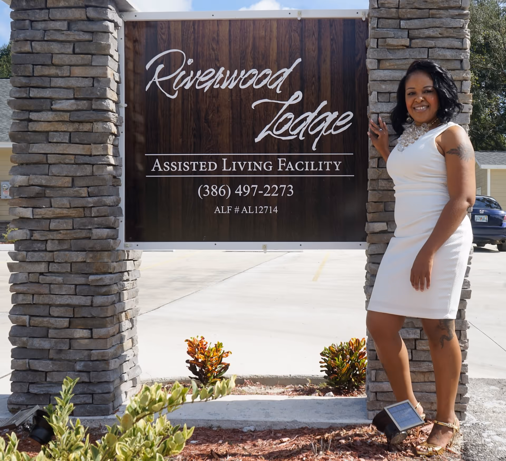 A woman in a white dress standing beside a stone sign that reads "Riverwood Lodge Assisted Living Facility."