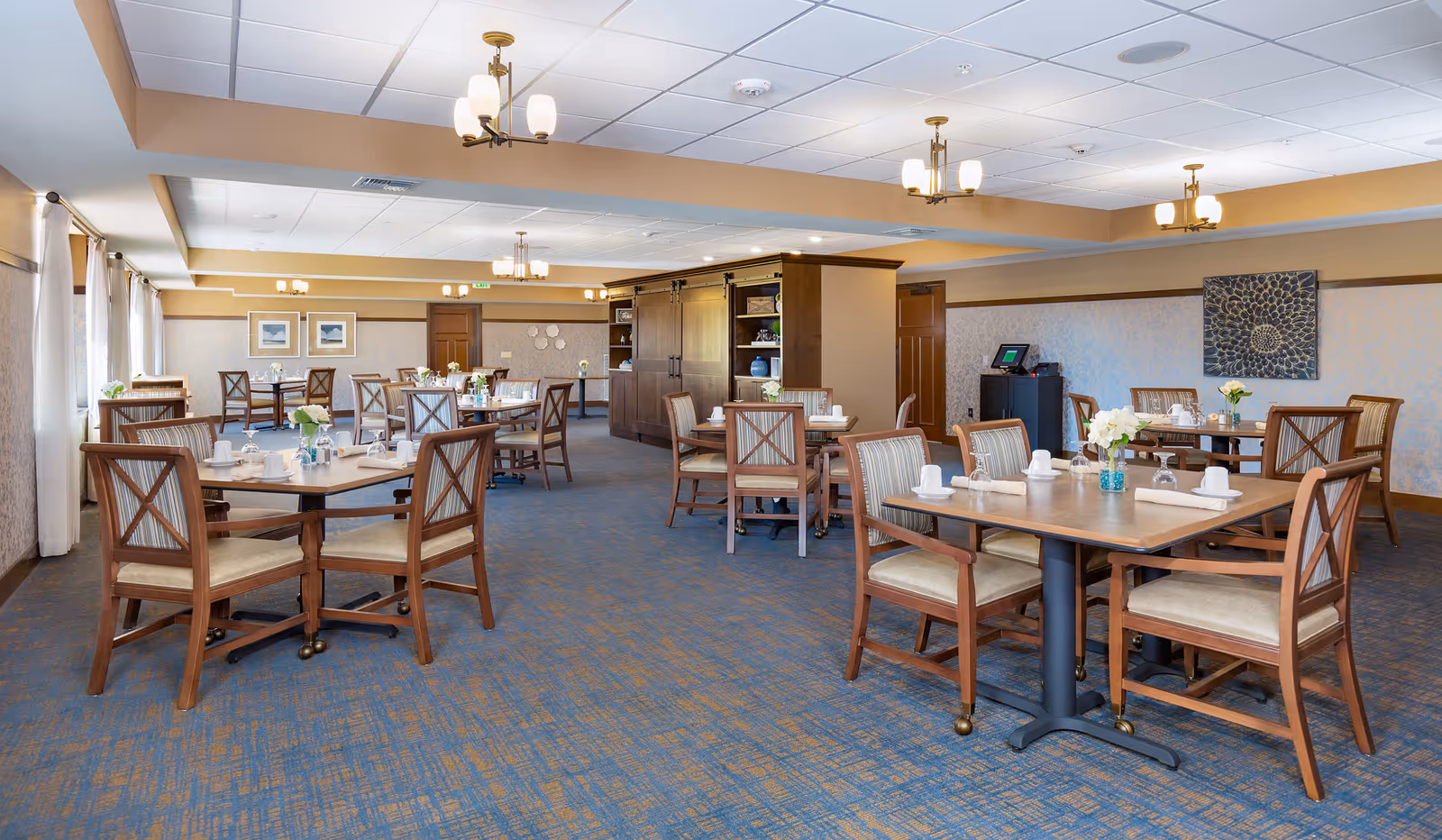 A spacious dining room with multiple wooden tables and chairs arranged neatly. Each table is set with white napkins, cups, and small flower vases. The room has blue and brown patterned carpet, beige walls with decorative artwork, and large windows with white curtains allowing natural light to enter. Ceiling lights with multiple bulbs provide additional illumination.