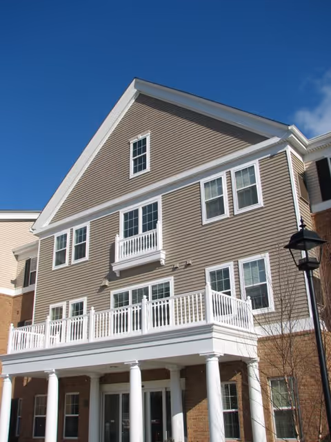 Exterior view of a multi-story senior living building with beige siding, white trim, multiple windows, a balcony with white railing, and white columns supporting a covered entrance under a clear blue sky.