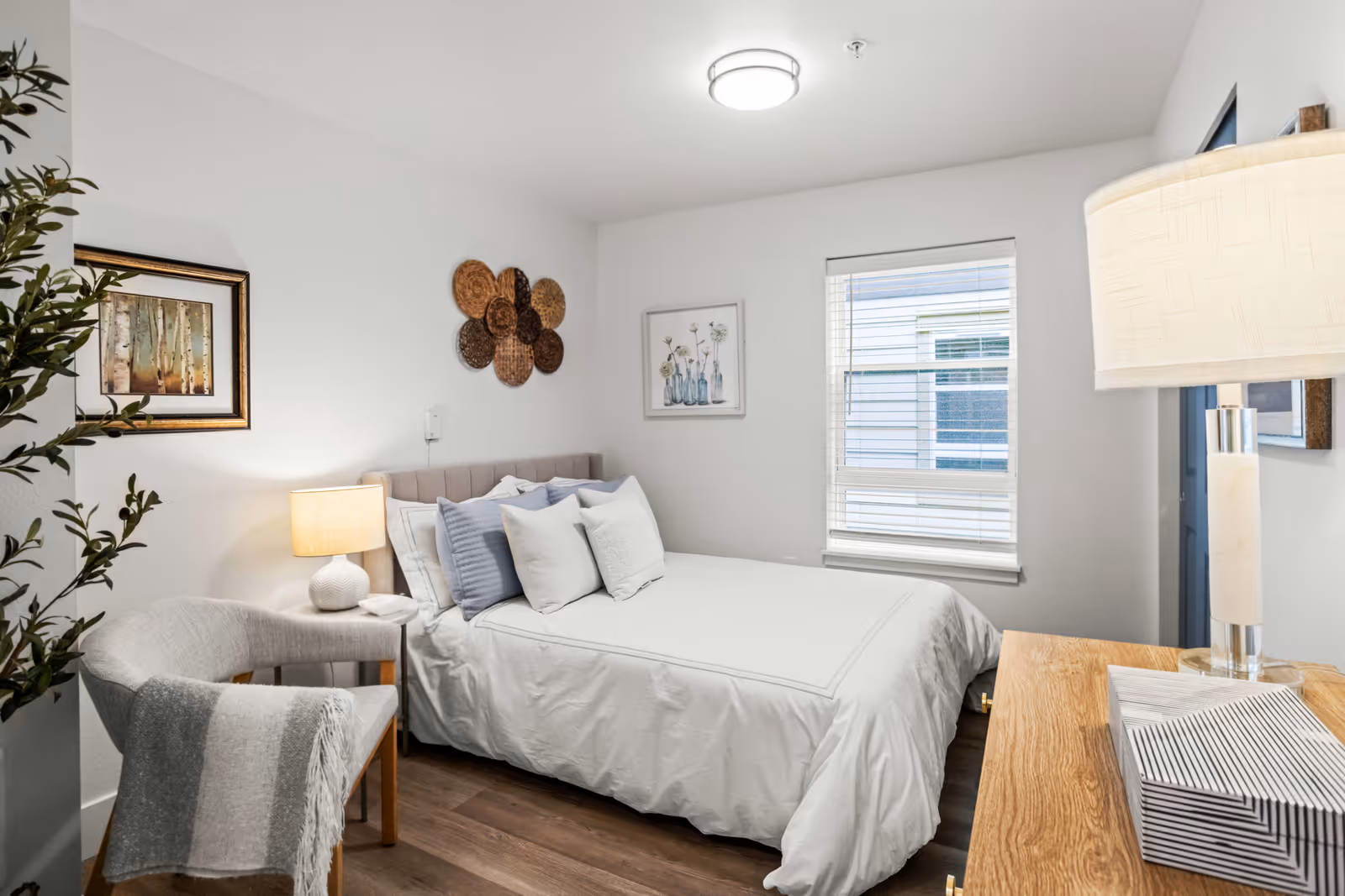 A cozy bedroom with a neatly made bed featuring white and light blue pillows. There is a wooden nightstand with a lamp on the left side of the bed, a gray armchair with a striped throw blanket, and a wooden dresser with a lamp on the right. The walls are decorated with framed artwork and woven wall hangings. A window with blinds lets in natural light.