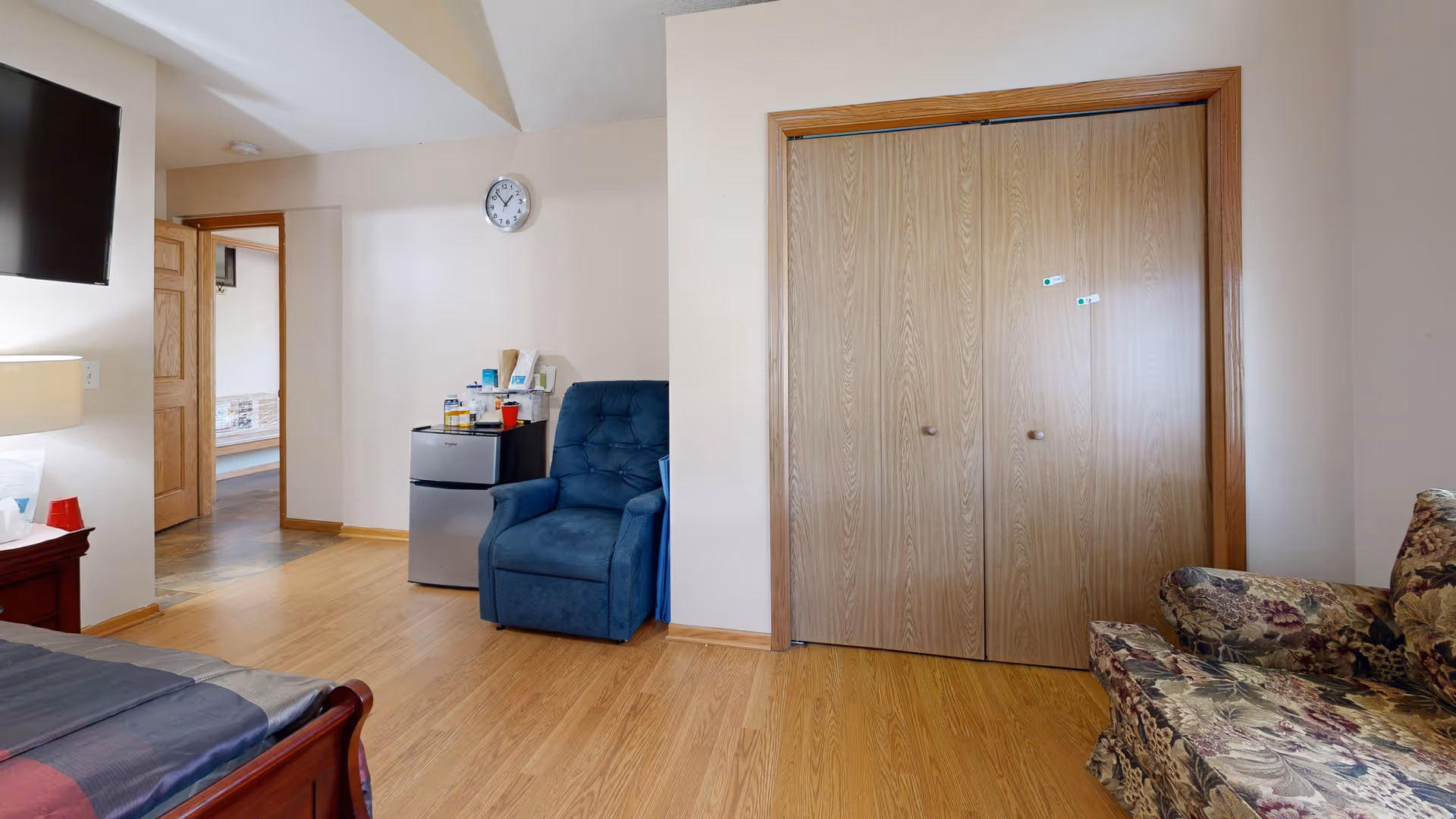 Interior view of a senior living facility room with wooden flooring, a blue armchair, a small refrigerator with various items on top, a floral patterned sofa, a wall clock, a wall-mounted TV, and a wooden closet with double doors.