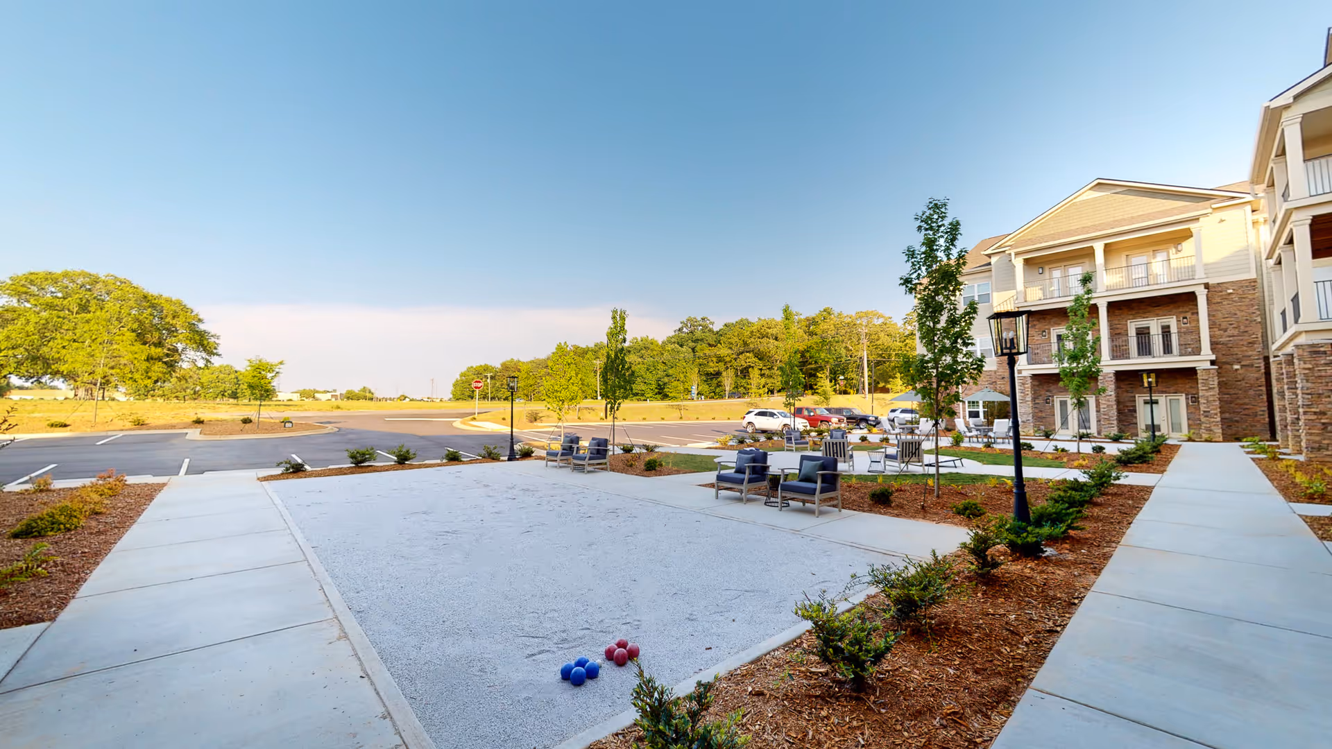 Outdoor courtyard area at Legacy Reserve at Fairview Park featuring a bocce ball court with balls, several seating areas with chairs and tables, landscaped plants, and a multi-story building with balconies in the background under a clear blue sky.