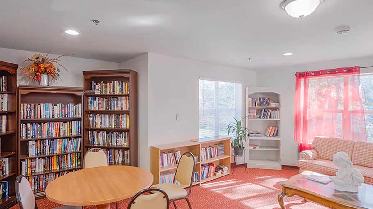 Sunlit common room with bookshelves, a round wooden table and chairs, and a sofa by windows with red curtains.