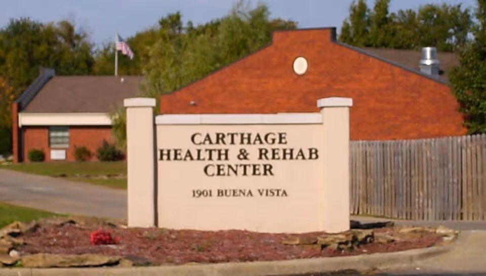 Outdoor view of the entrance sign for Carthage Health & Rehab Center located at 1901 Buena Vista, with a brick building, a wooden fence, and trees in the background.
