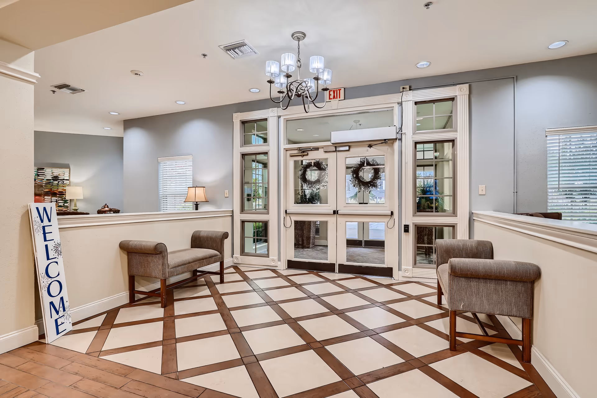 Entrance area of a facility with double glass doors decorated with wreaths, two cushioned benches on either side, a chandelier hanging from the ceiling, a 'WELCOME' sign leaning against the wall, and windows with blinds allowing natural light.