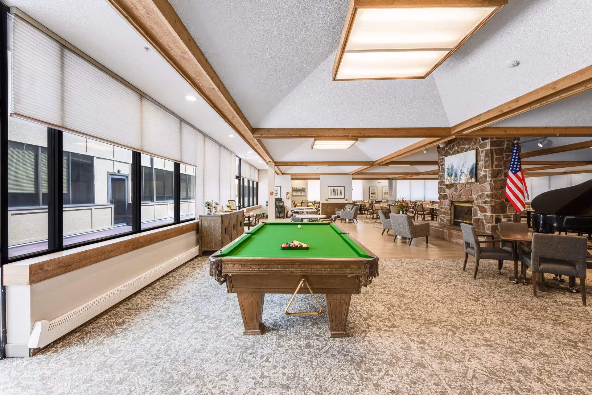Spacious senior living common room with a pool table centered, seating areas, a stone fireplace, a piano, and large windows.