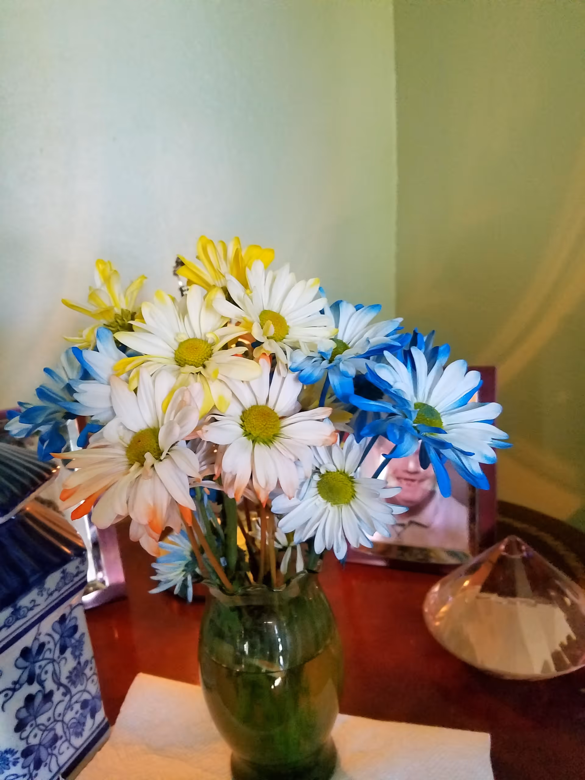 A bouquet of white daisies with yellow centers, some petals dyed blue and orange, arranged in a green glass vase on a wooden surface. In the background, there is a framed photo of a person and a decorative glass object.