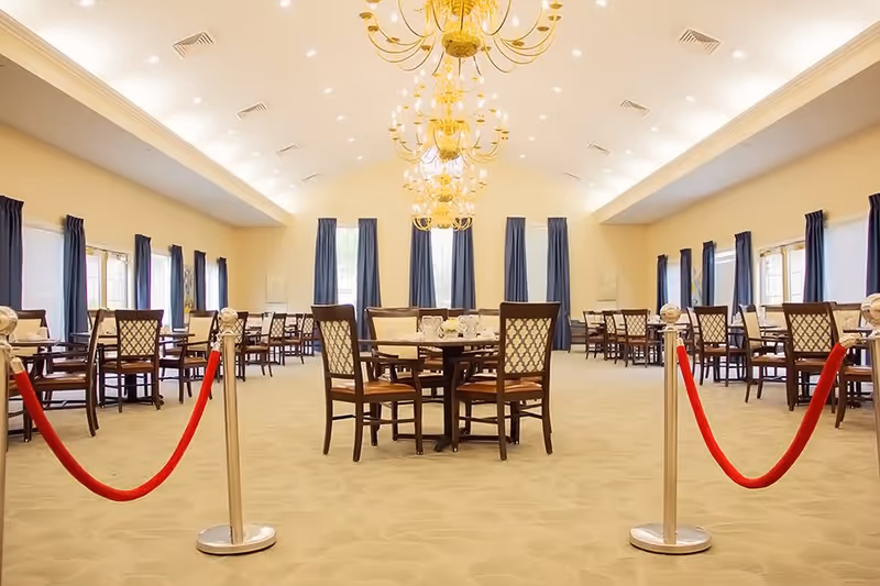 A spacious dining room with multiple round tables and wooden chairs arranged neatly. The room features large windows with dark blue curtains, beige walls, and a high ceiling adorned with elegant gold chandeliers. Red velvet ropes on silver stanchions block off the area in the foreground.