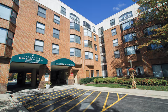 Exterior view of a multi-story brick retirement facility building with green awnings over the entrances labeled 'Broaddale Hawthorn Lakes' and '10 East Hawthorn Drive'. There is a parking area with yellow lines and some landscaping with bushes and a tree on the right side under a clear blue sky.