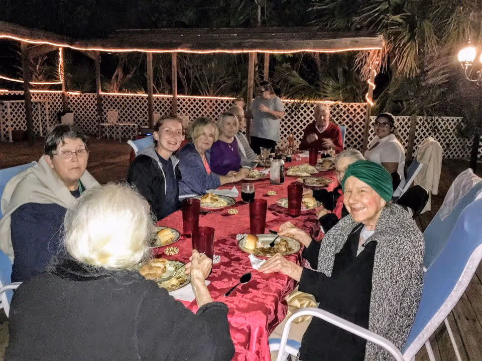 A group of elderly people and caregivers sitting around a long table outdoors at night, enjoying a meal together. The table is covered with a red tablecloth and set with plates of food and red cups. There are string lights and a white lattice fence in the background, creating a cozy atmosphere.