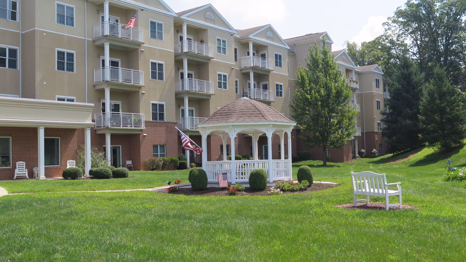 Outdoor view of a senior living facility with a multi-story building in the background, featuring balconies and windows. In the foreground, there is a white gazebo surrounded by small bushes and flowers, an American flag, and a white bench on a well-maintained green lawn with trees.