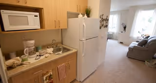 A small kitchen area with wooden cabinets, a microwave, coffee maker, sink, and refrigerator. The kitchen opens into a carpeted living room with a sofa and large windows letting in natural light.