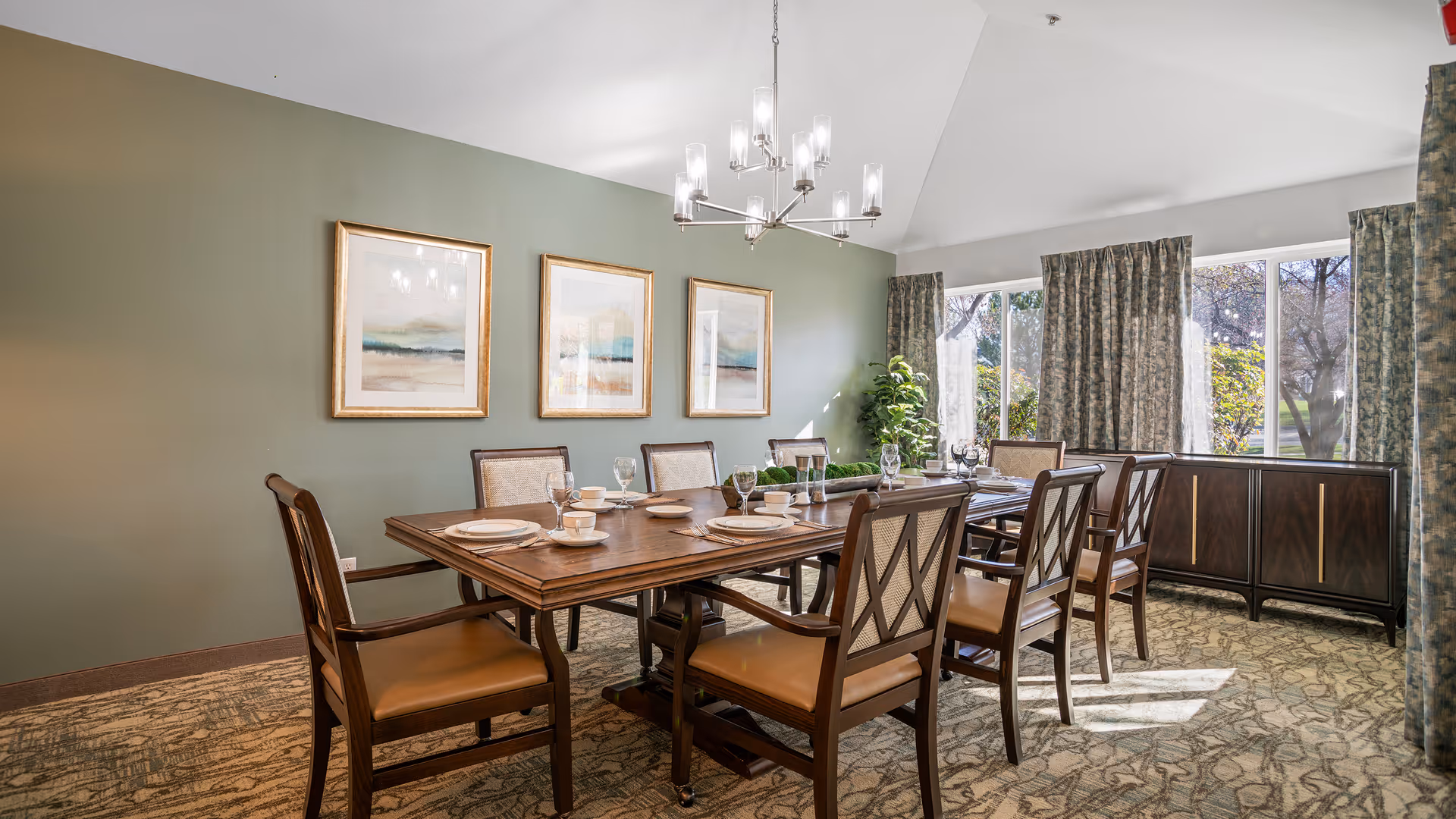 A well-lit dining room with a large wooden dining table set for eight people. The table has plates, cups, and glasses arranged neatly. The room features a green accent wall with three framed landscape paintings, a modern chandelier hanging from the ceiling, patterned carpet, and large windows with curtains letting in natural light. A dark wooden sideboard and a potted plant are visible near the windows.
