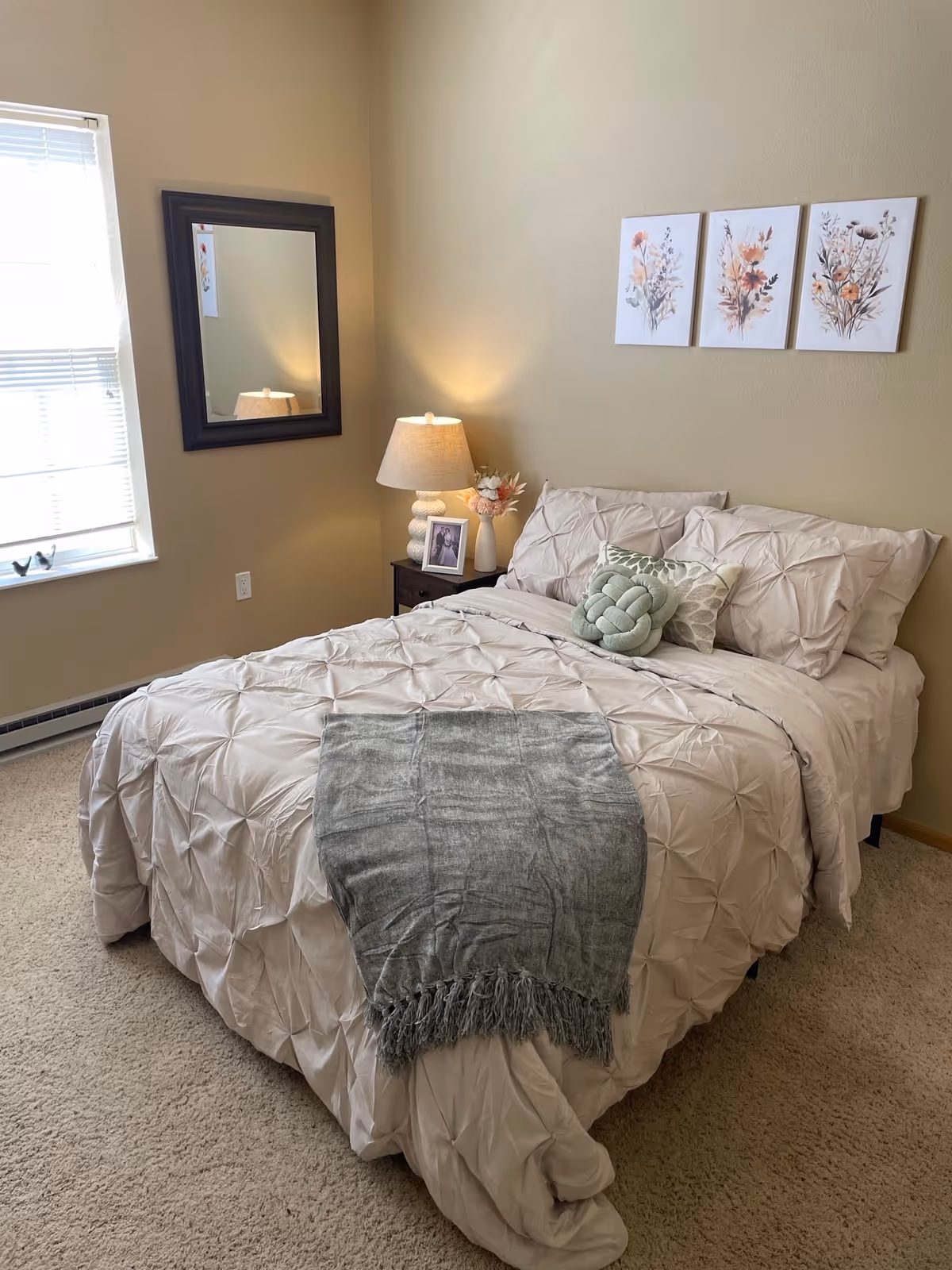 A cozy bedroom with a neatly made bed featuring light beige bedding, two decorative pillows, and a gray throw blanket at the foot. A nightstand beside the bed holds a lamp, a framed photo, and a small vase with flowers. Above the bed, there are three floral-themed wall art pieces. A window with blinds is on the left wall, and a large dark-framed mirror hangs next to it.