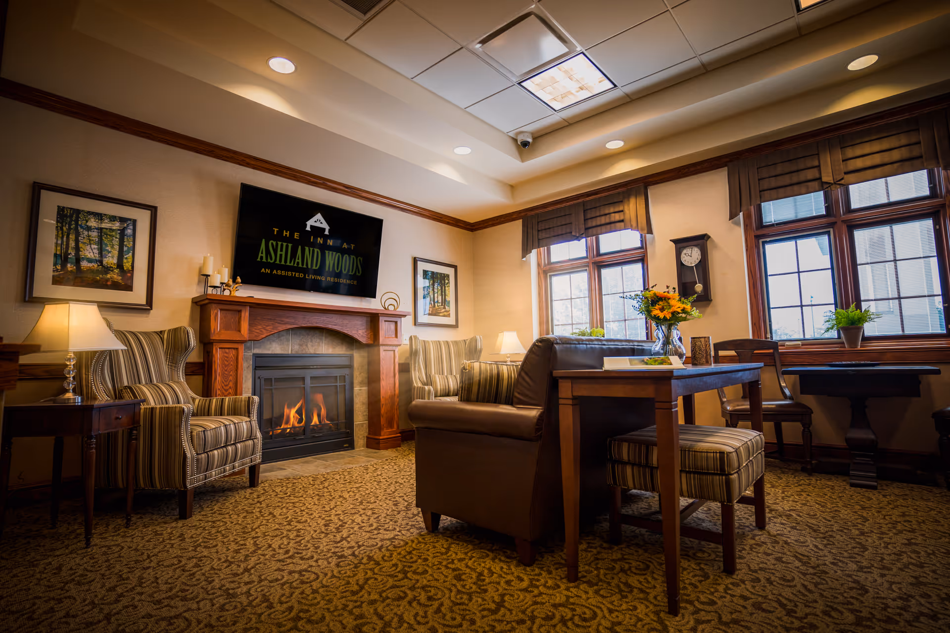 A cozy living room area in The Inn at Ashland Woods featuring a lit fireplace with a wooden mantle, a flat-screen TV mounted above displaying the facility's name, two striped armchairs, a brown leather sofa, a wooden table with a vase of flowers, and large windows with brown valances letting in natural light.