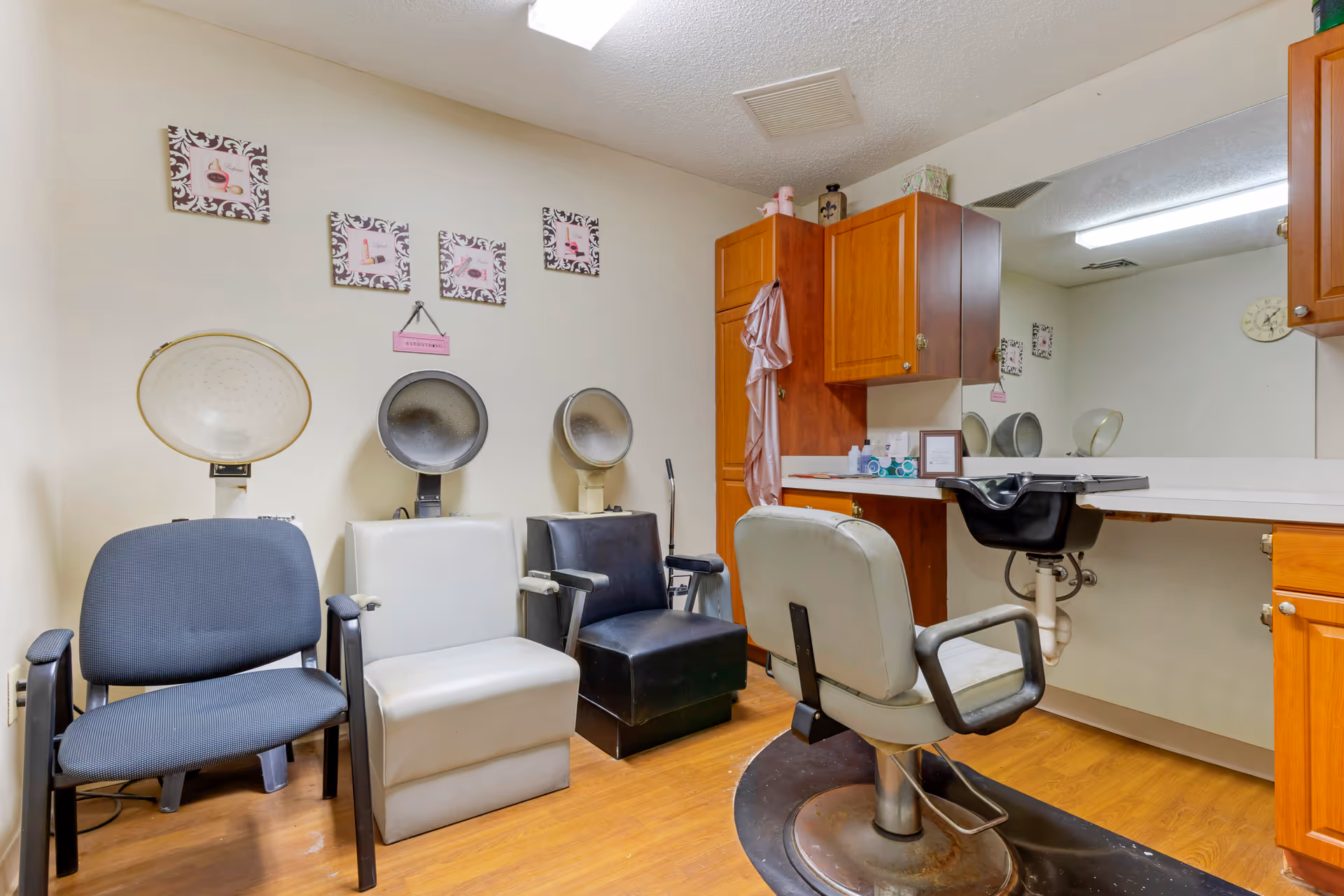 Interior of a salon area in a senior living facility with three vintage hair drying chairs lined up against a wall and a barber chair in front of a large mirror. Wooden cabinets and a countertop with a sink are visible on the right side. The walls are decorated with framed pictures and a small hanging sign.