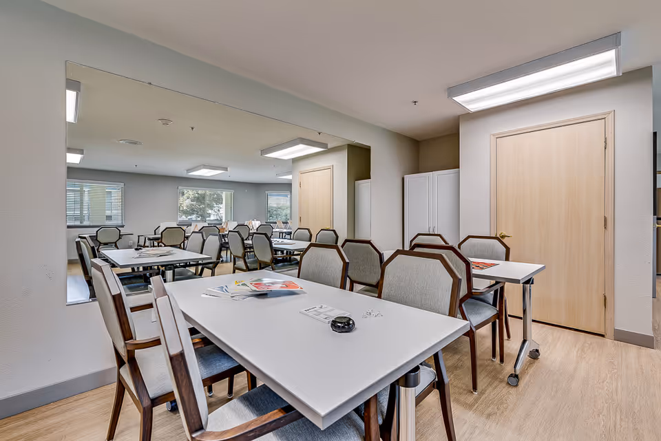 A bright, clean room with multiple tables and chairs arranged for group activities or dining. The room has light-colored walls, large windows with blinds, and a large mirror on one wall reflecting the tables and chairs. There are some papers and small items on the tables, and a closed wooden door is visible in the background.