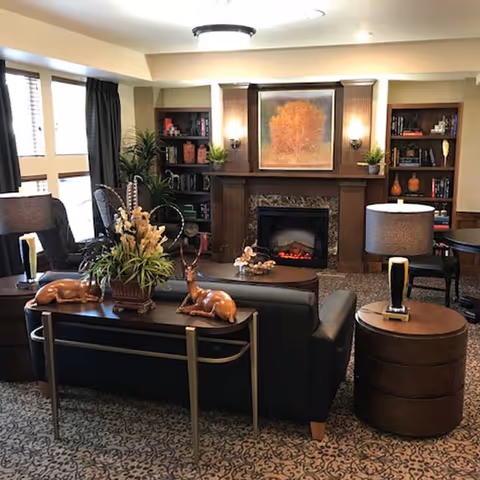 Cozy common living room with a fireplace, sofa, bookshelves, lamps, and decorative plants.
