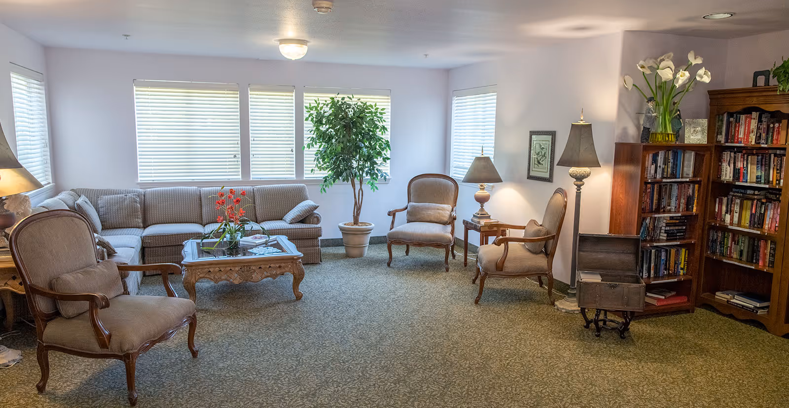 A cozy communal living room with a sectional sofa, several armchairs, a coffee table, potted plant, lamps, and a bookshelf.