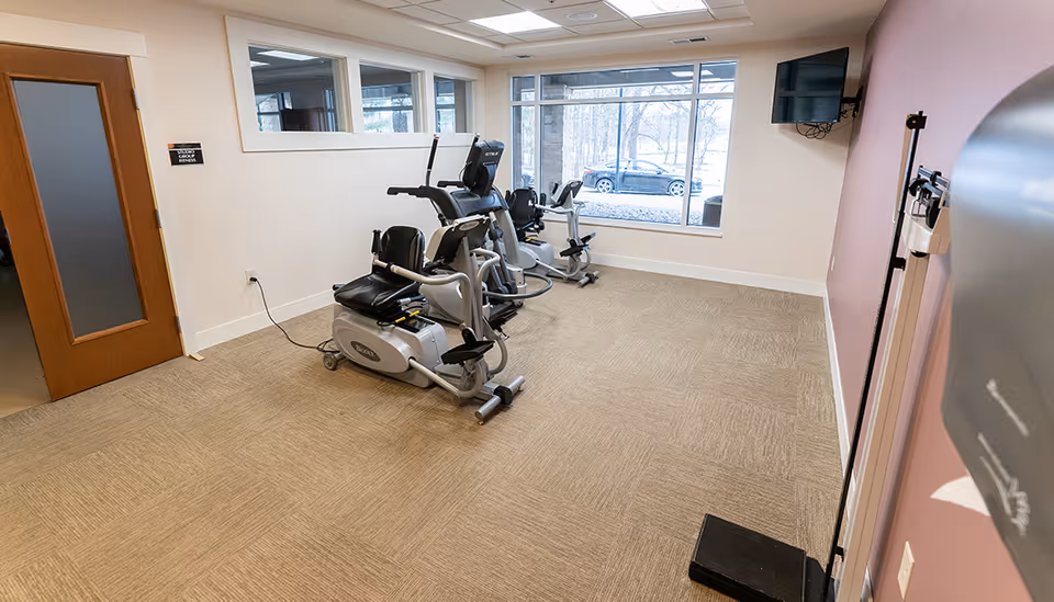 A small fitness room with three exercise bikes lined up near a large window showing a view of parked cars outside. The room has beige carpet, white walls, a wooden door, a wall-mounted TV, and a scale in the corner.