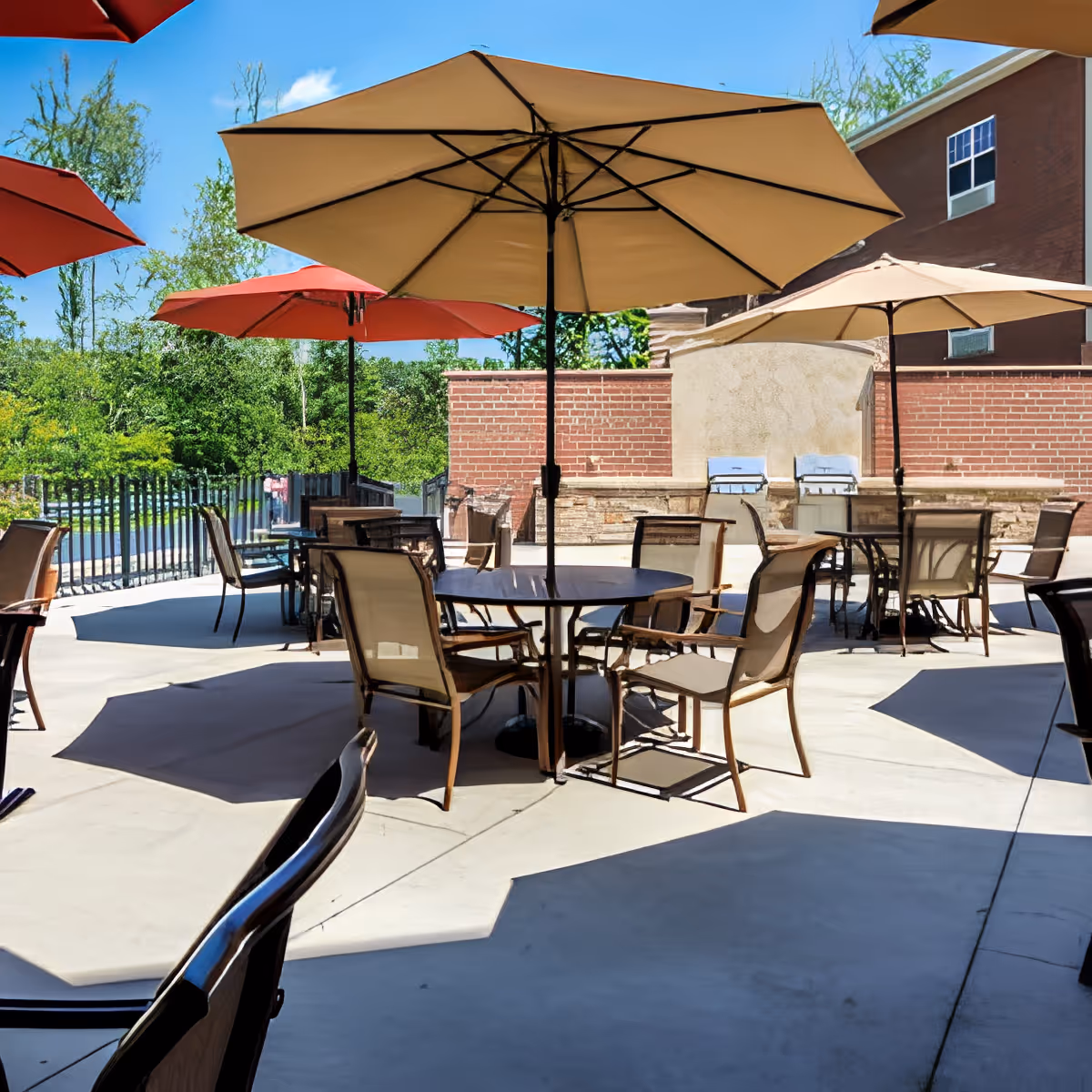 Outdoor patio area with multiple round tables and chairs under large beige and red umbrellas, surrounded by greenery and a brick wall with two built-in grills.