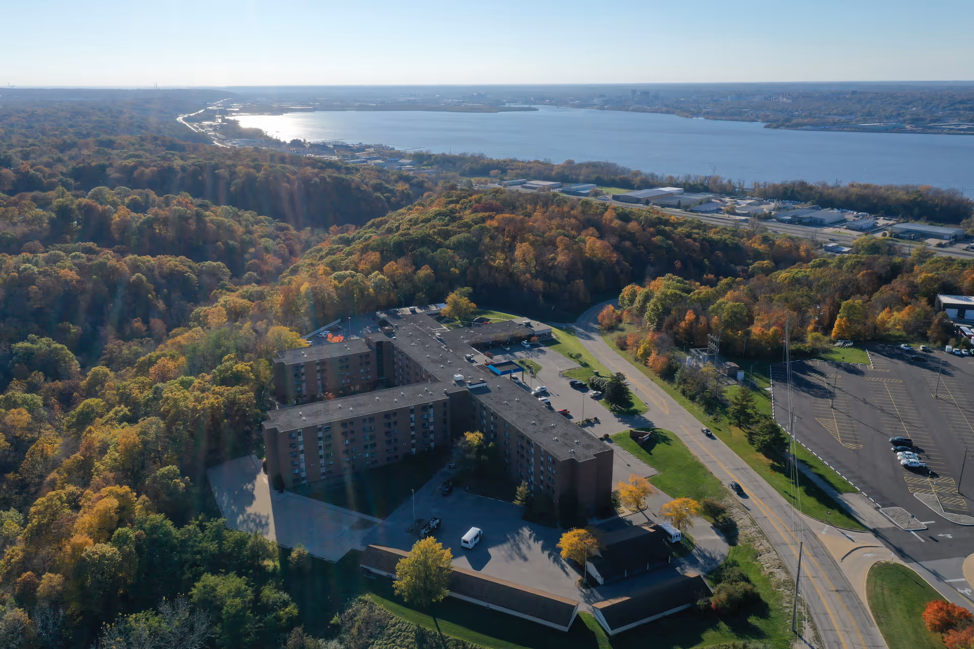 Aerial view of Saxony Court Senior Living facility surrounded by dense trees with autumn foliage, a large parking lot, and a river in the background under a clear sky.