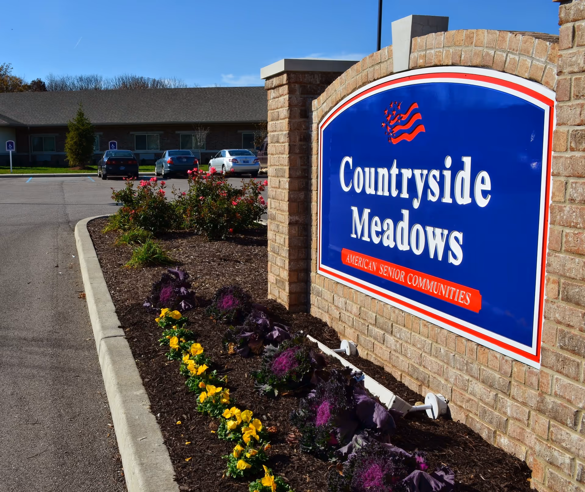 A large blue and red sign reading 'Countryside Meadows American Senior Communities' mounted on a brick wall next to a flower bed with yellow and purple flowers. In the background, there is a parking lot with several cars and a single-story building under a clear blue sky.
