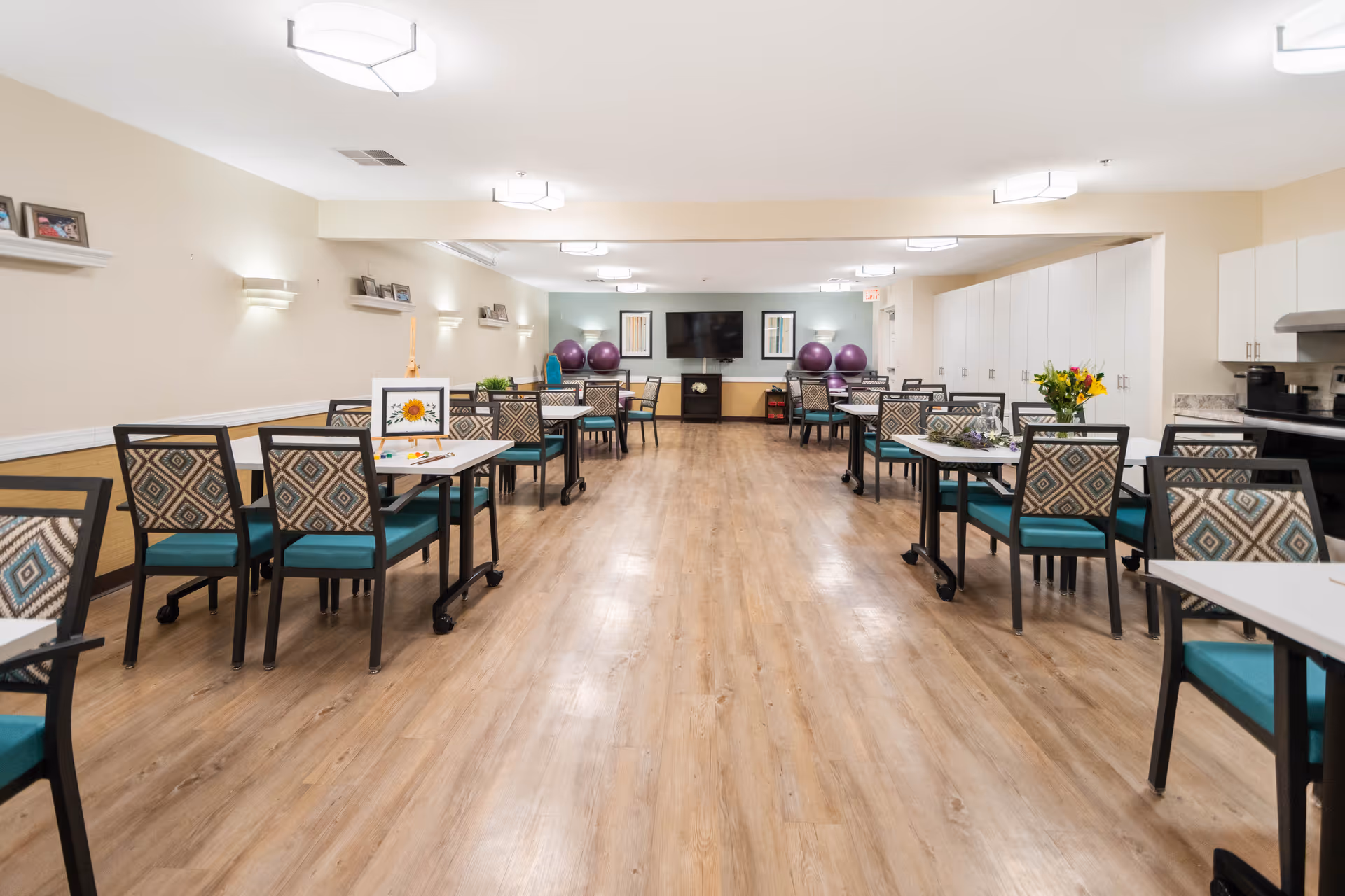 A spacious, well-lit common room with multiple tables and chairs arranged neatly. The chairs have patterned backs and teal seats. The room has light-colored walls with small shelves holding framed photos and wall-mounted lights. At the far end, there is a large flat-screen TV mounted on the wall with exercise balls and framed artwork nearby. The floor is wood-style laminate, and there are cabinets and a kitchenette area on the right side.
