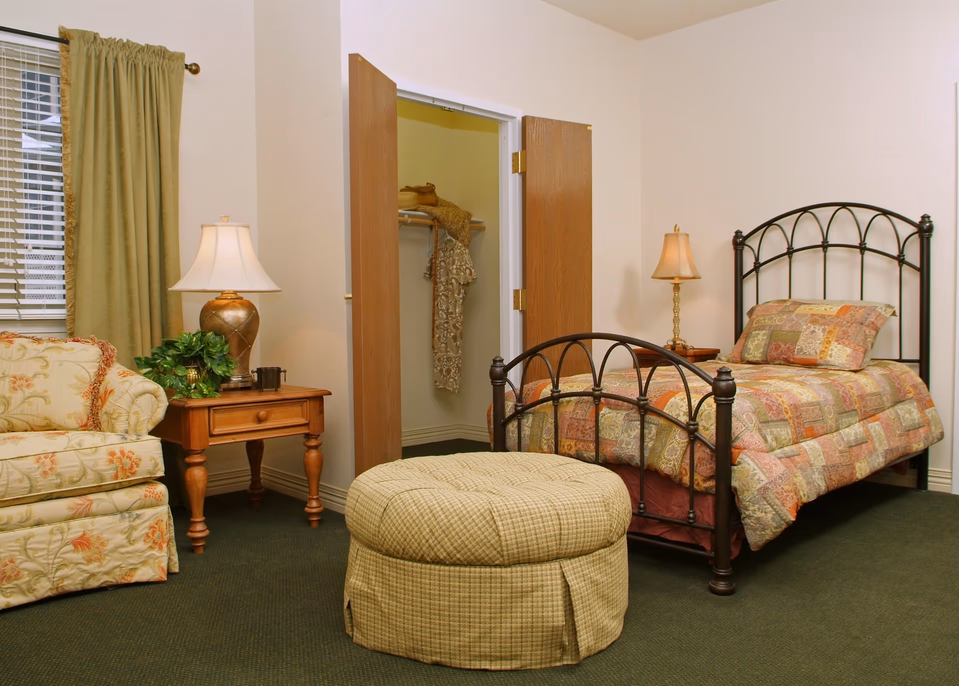 A furnished bedroom with a single iron-frame bed, patterned bedding, a sofa, ottoman, and bedside tables with lamps.
