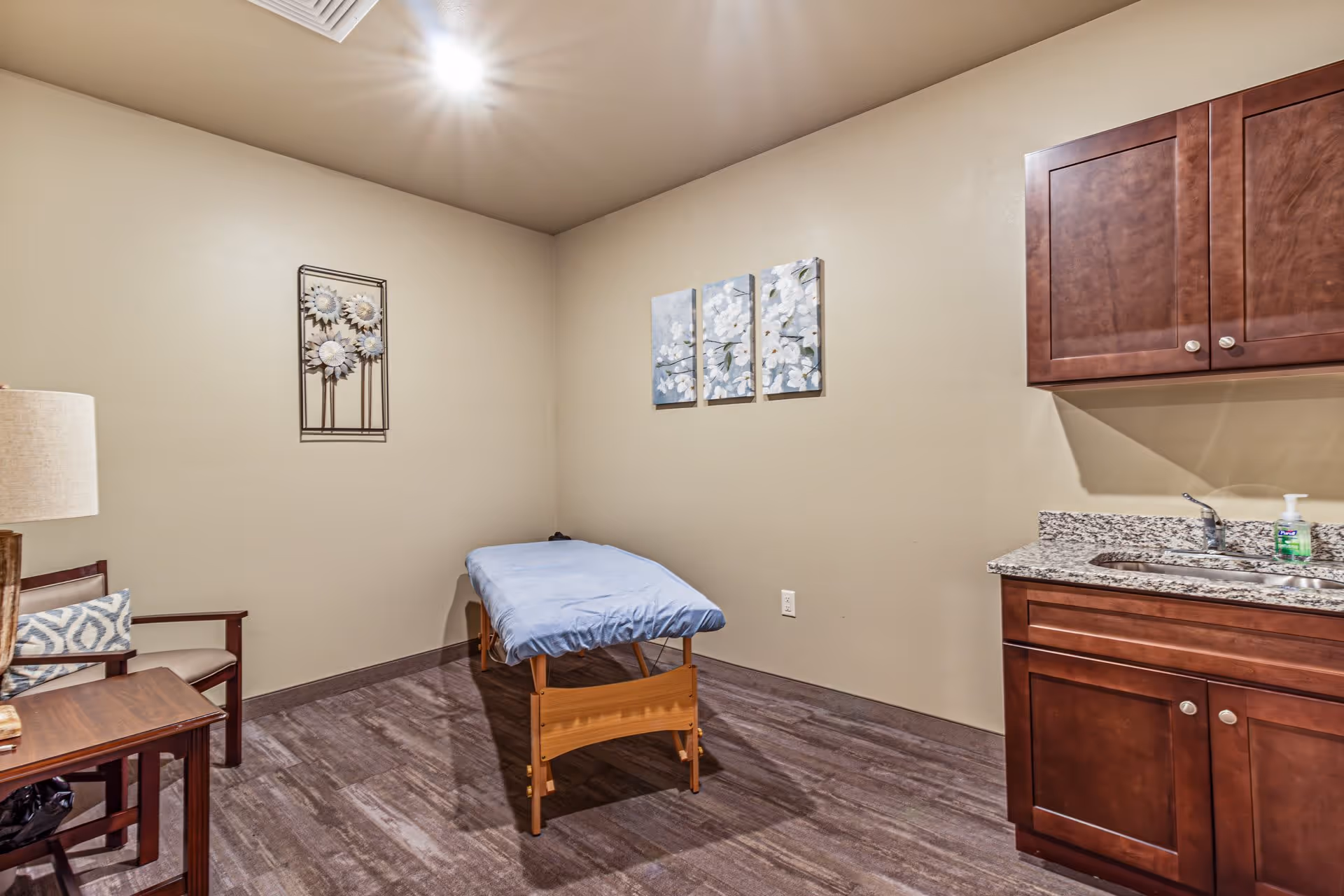 A small room with a massage or treatment table covered with a blue sheet, a wooden chair with a cushion, a wooden side table with a lamp, and a countertop with a sink and cabinets. The walls are beige with decorative wall art featuring flowers.
