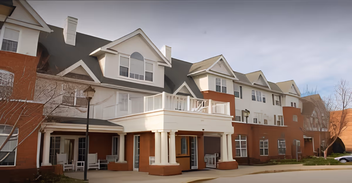 Exterior view of a multi-story senior living facility building with a combination of red brick and white siding, featuring multiple windows, a covered entrance supported by columns, and a small balcony above the entrance.