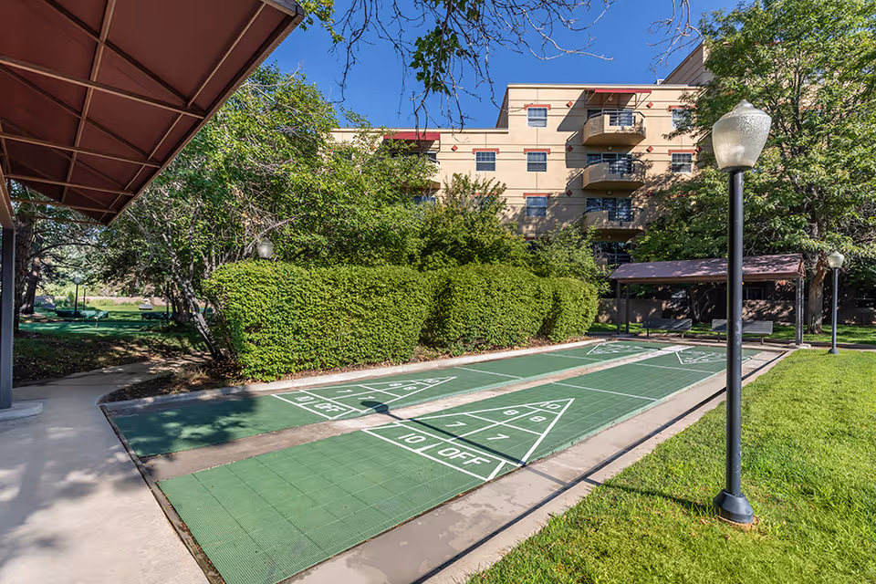 Outdoor shuffleboard courts with green playing surfaces and white markings, surrounded by green grass, bushes, and trees. A multi-story beige building with balconies is visible in the background under a clear blue sky. There are lamp posts and a covered seating area near the courts.