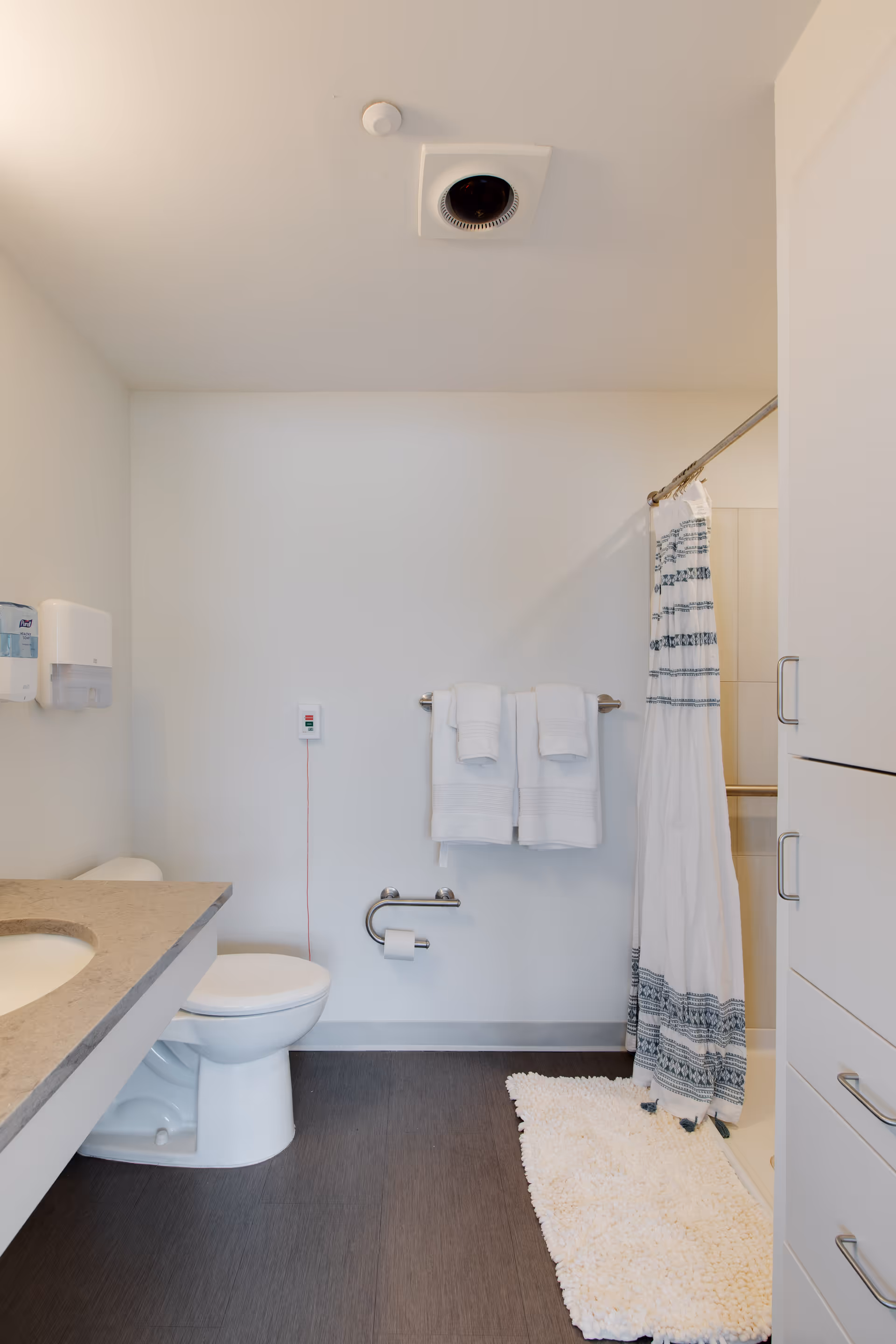 Clean, accessible bathroom with a toilet, countertop sink, towel rack, shower with a patterned curtain and a white bath mat.