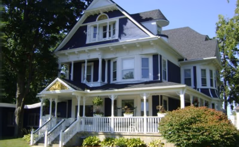 A large, two-story Victorian-style house painted dark blue with white trim. It features a wrap-around porch with white railings and columns, multiple windows including a dormer window on the roof, and is surrounded by greenery including bushes and trees under a clear blue sky.