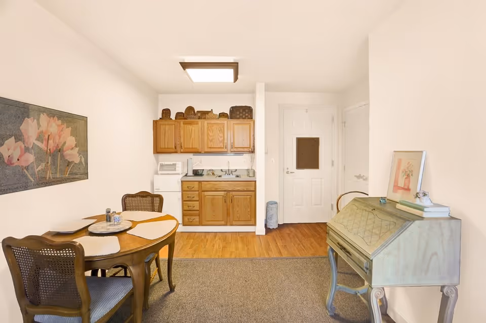 A small dining area and kitchenette in a senior living facility. The room features a wooden dining table with four chairs, placemats, and a centerpiece. The kitchenette has wooden cabinets, a small sink, a microwave, and a mini refrigerator. There is a decorative painting on the wall to the left and a vintage-style desk with books and a framed picture on the right. The floor is a combination of carpet and wood, and the walls are painted white.