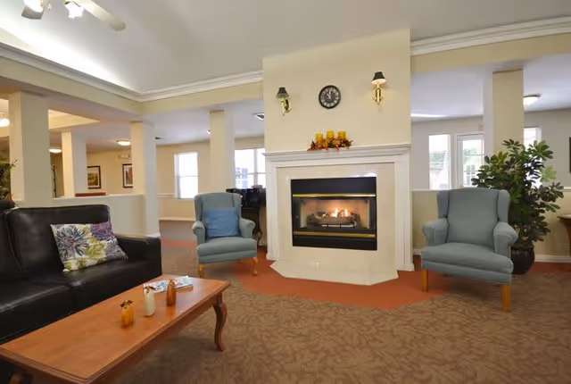 A cozy senior living facility common area with a lit fireplace in the center. Two blue armchairs are placed on either side of the fireplace, and a black leather couch with a floral pillow is on the left. A wooden coffee table with small decorative items is in front of the couch. The room has beige walls, carpeted floors, and large windows letting in natural light.