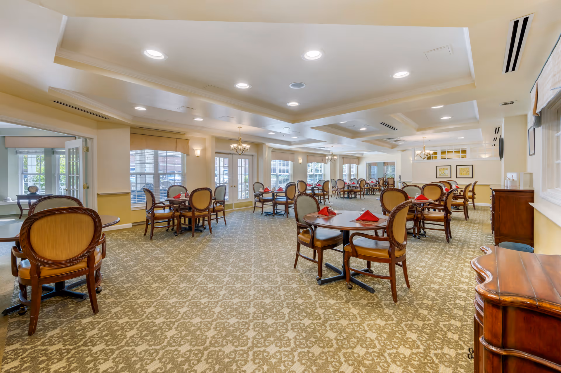 A spacious dining room with multiple round tables and wooden chairs with cushioned seats. Each table is set with red folded napkins. The room has large windows allowing natural light, beige walls, patterned carpet, and ceiling lights with chandeliers.