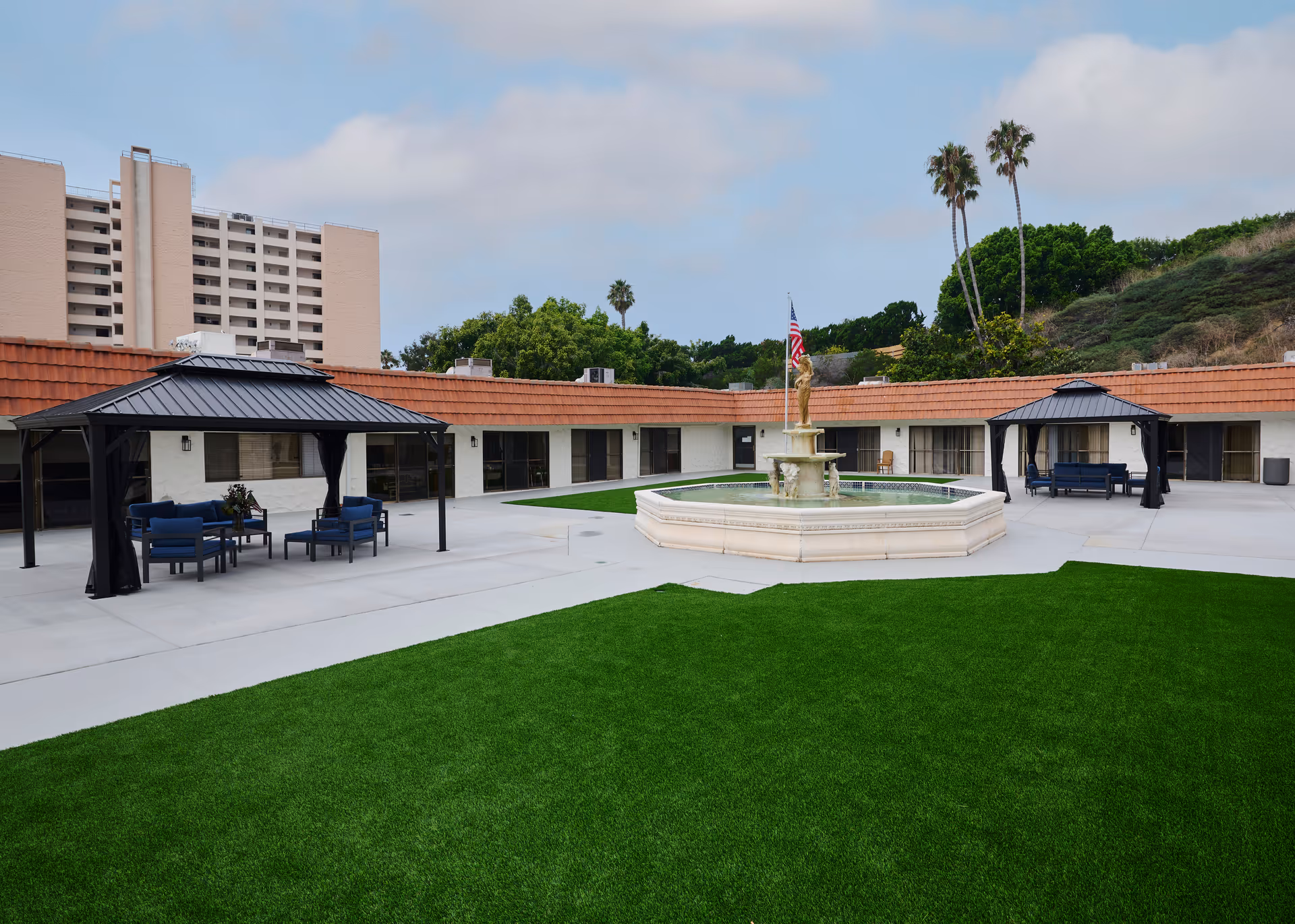 Outdoor courtyard area of a nursing and rehabilitation center featuring a large octagonal fountain with a statue in the center, surrounded by a concrete patio and green artificial turf. There are two black metal gazebos with blue cushioned seating and tables. In the background, there are single-story buildings with red-tiled roofs and a taller building visible behind them. Palm trees and other greenery are also visible.