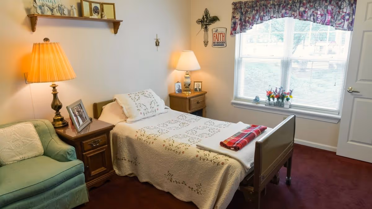 A cozy bedroom with a single bed covered in a white floral quilt and a red plaid blanket folded at the foot. There are two wooden nightstands on either side of the bed, each with a lamp and framed photos. A green armchair with a white cushion is next to one nightstand. A window with floral valance lets in natural light, and a decorative cross and sign are on the wall.